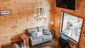 A cozy log cabin interior with a sofa, two wooden coffee tables, a chandelier, and a large window with a view. - Stunning Hocking Hills Views, Huge Skies ~ Sauna, Pet Friendly, Hot Tub, WiFi (new plymouth)