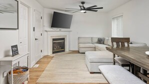 A modern living room with a fireplace, a television, a sofa, a dining table, and a ceiling fan. - Wonderfully Updated, Pet Friendly, Open Layout, Pool Table, BBQ, Next to Edgefield Golf and Concerts (Troutdale)