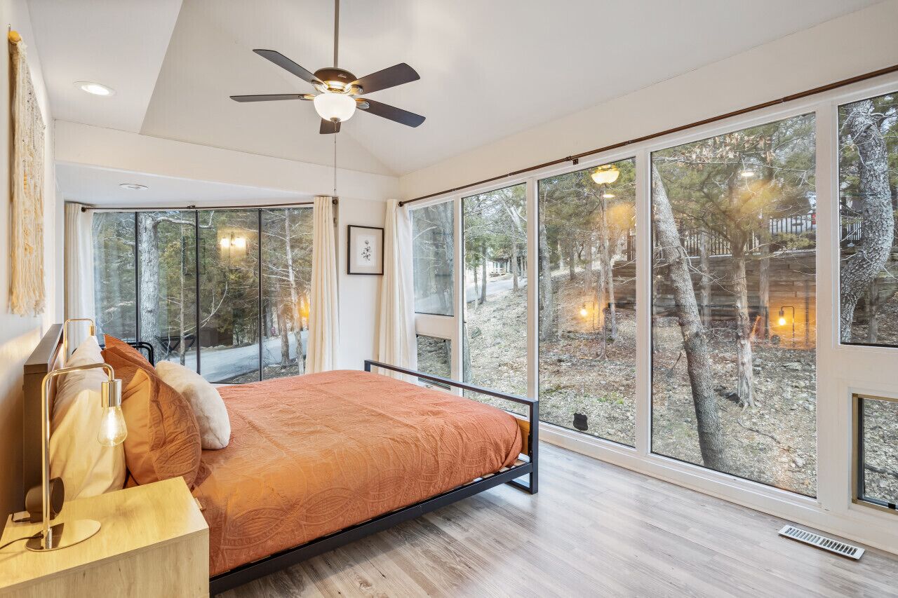 Un dormitorio con cama, ventilador de techo, ventanas amplias y vista a árboles.