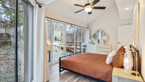 A bedroom with a bed, a ceiling fan, a dresser, a mirror, and large windows. - Tree Top Hideaway – Pools, Sauna & More! (Lake Ozark)