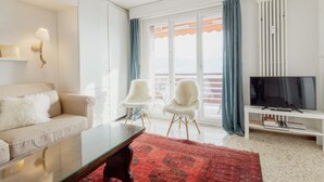 Ein modernes Wohnzimmer mit einer Couch, zwei Sesseln, einem Fernseher und einem großen Fenster mit Vorhängen.