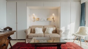 Ein Wohnzimmer mit einem beigen Sofa, einem Glastisch, einem roten Teppich mit Muster, einem Holztisch und einem weißen Stuhl.