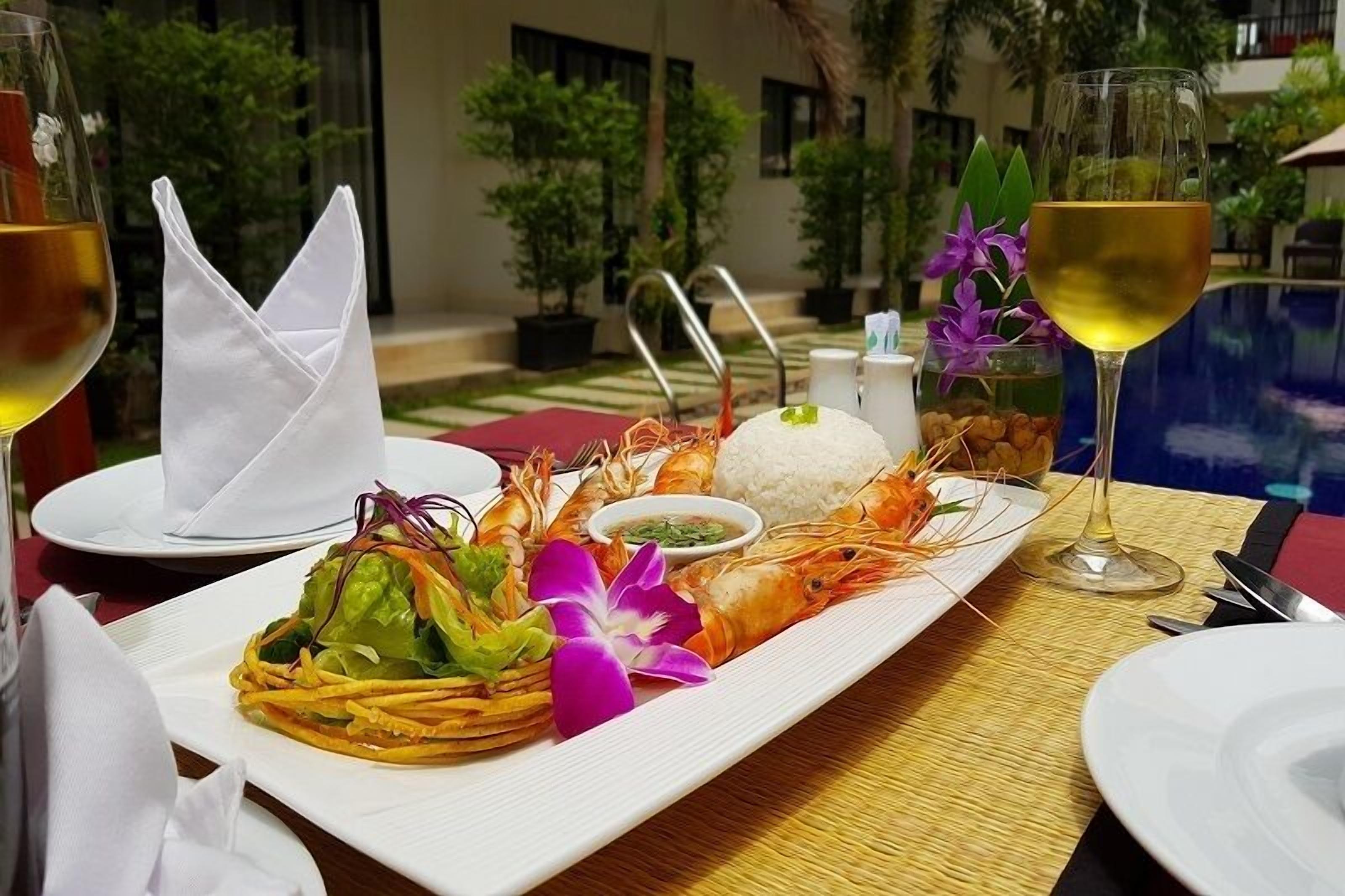 Una mesa de comedor con un plato de mariscos, un bol de arroz y dos copas de vino blanco, con vista a una piscina y follaje.