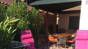 An outdoor patio with two pink chairs, a wooden table, and a covered area with a tiled roof. - Rent House in Residence (Argelès-sur-Mer)