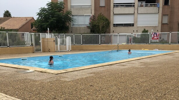 A swimming pool with a tiled deck area, surrounded by a white fence and a building with multiple balconies. - Rent House in Residence (Argelès-sur-Mer)