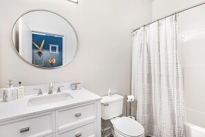 A modern bathroom with a white sink, vanity, and toilet. A round mirror hangs above the sink, and a shower curtain is visible. - Walker's Paradise + Espresso Machine + King Bed (Atlanta)
