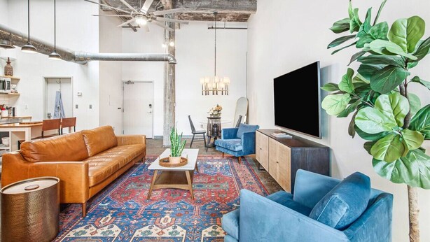 A living room with a brown leather sofa, blue armchairs, a coffee table, and a large flat-screen TV. There is a patterned rug on the floor and a plant on the right side. - Walker's Paradise + Espresso Machine + King Bed (Atlanta)