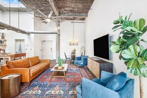 A living room with a brown leather sofa, blue armchairs, a coffee table, and a large flat-screen TV. There is a patterned rug on the floor and a plant on the right side. - Walker's Paradise + Espresso Machine + King Bed (Atlanta)