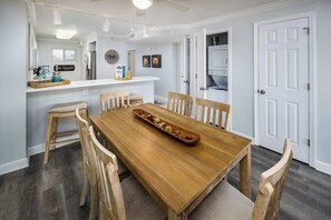 A dining area with a wooden table and chairs, a kitchen counter with a refrigerator, and a door leading to another room.