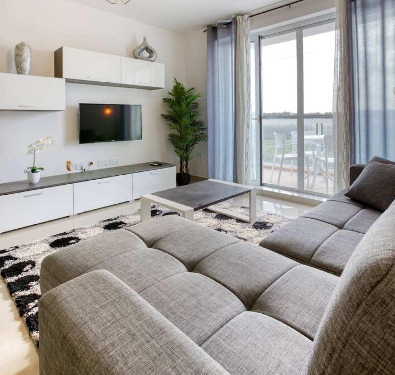 Un salon moderne avec un canapé gris, un téléviseur à écran plat fixé au-dessus d’une console blanche et une grande fenêtre agrémentée de rideaux.