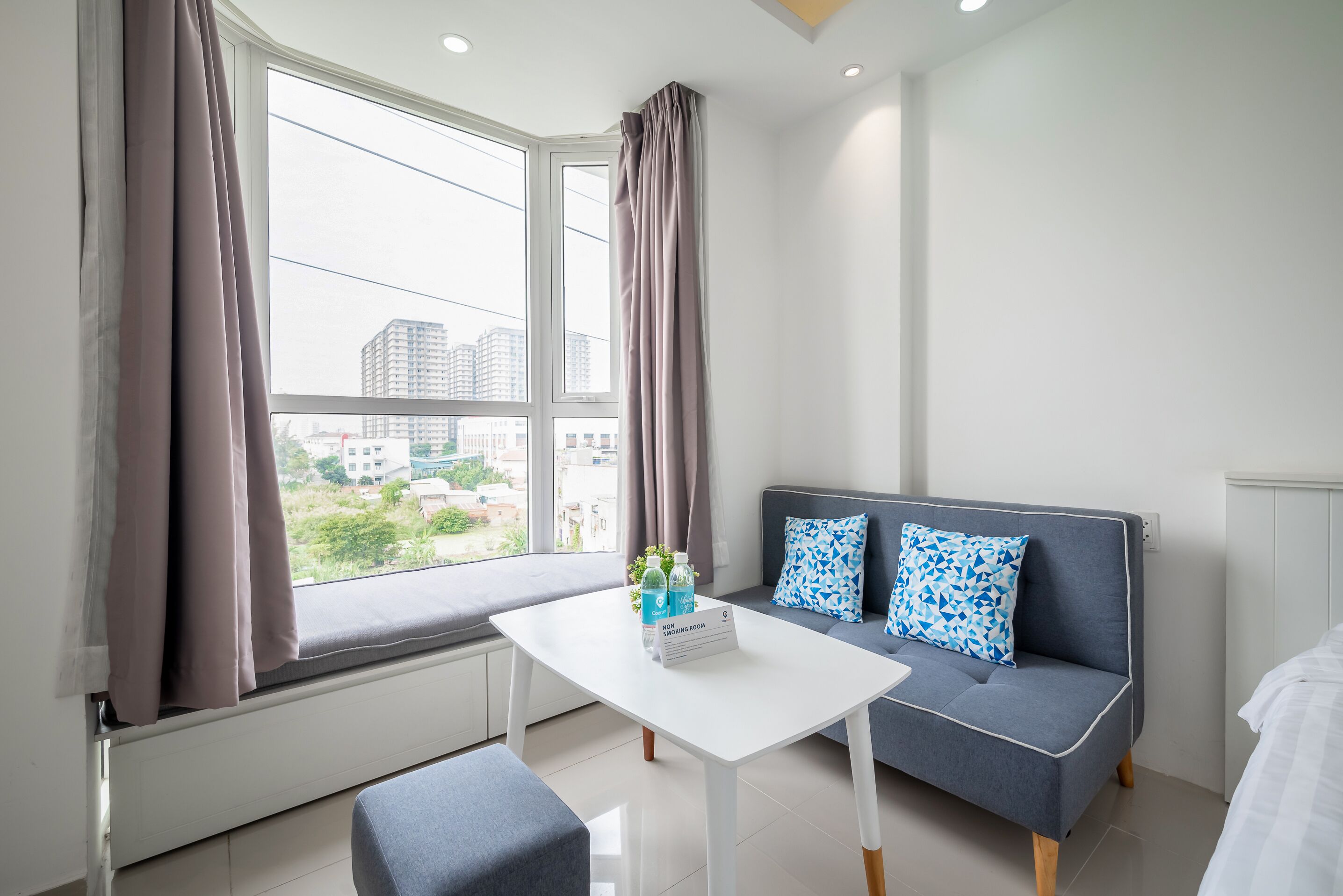 Een moderne kamer met een witte tafel, een grijze bank en grote ramen voorzien van gordijnen.