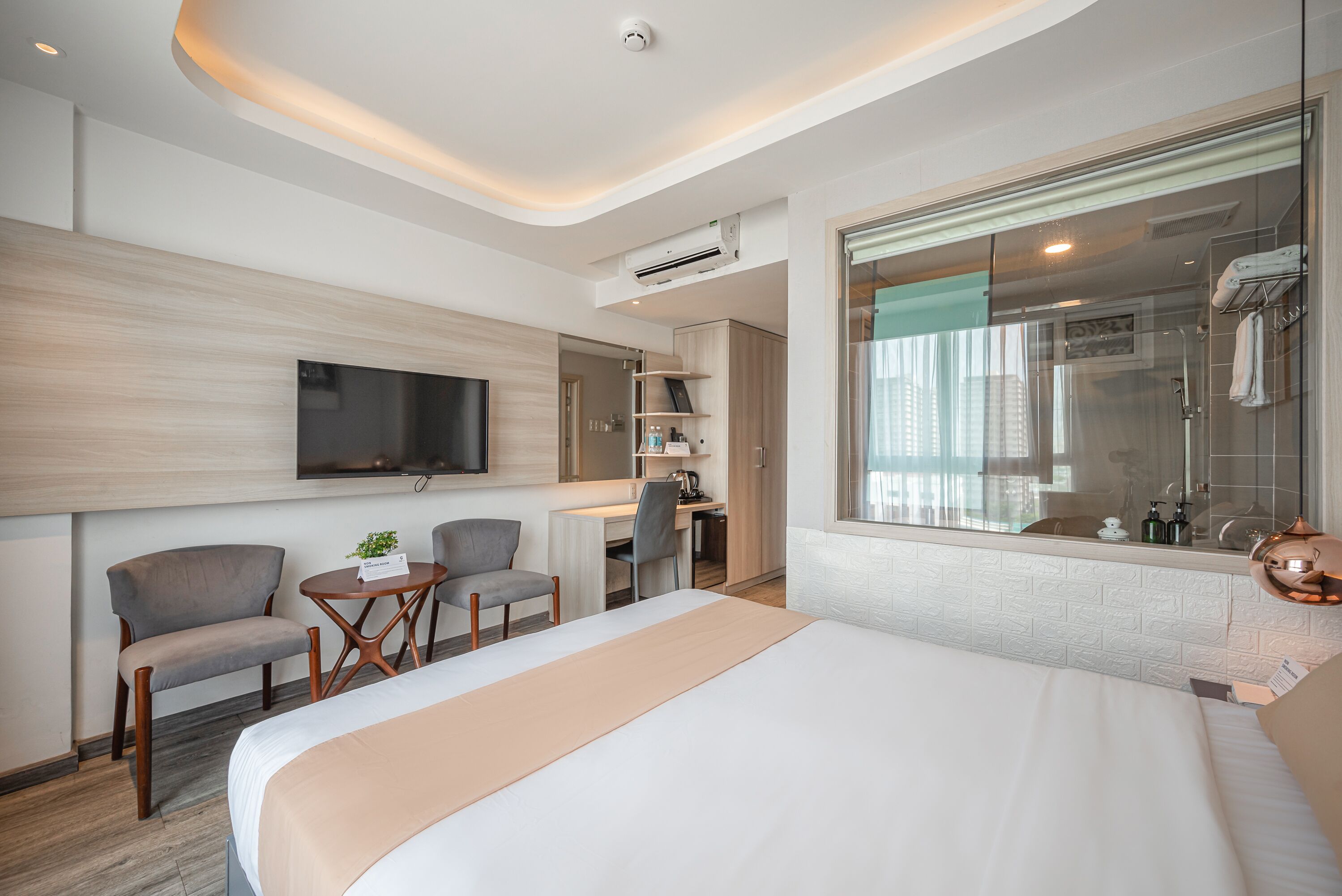 A modern hotel room with a large bed, a desk with a chair, a TV mounted on the wall, and a shower area with a glass door.