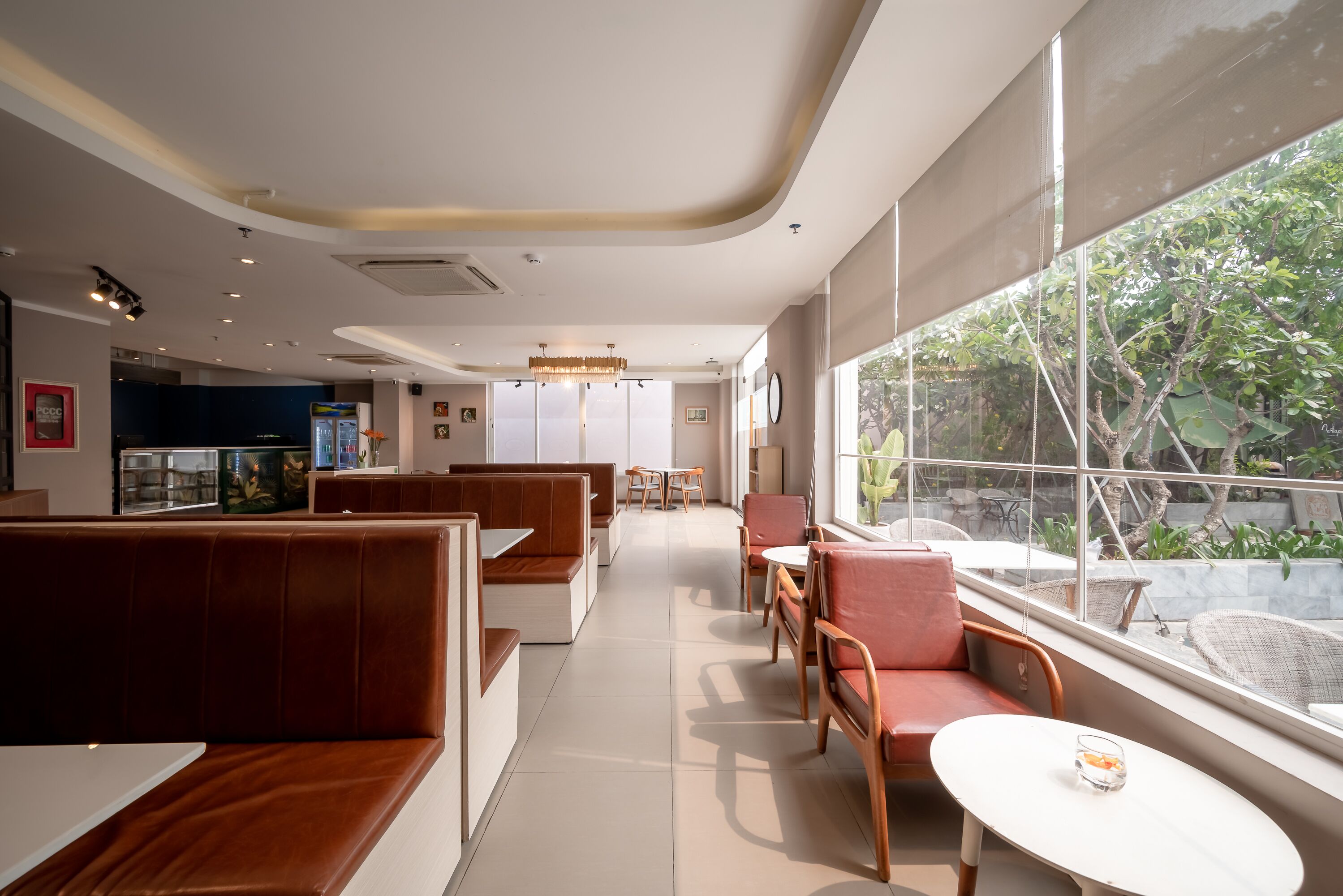 Een modern restaurant met zitjes en tafels, grote ramen met uitzicht op groen, en een kroonluchter.