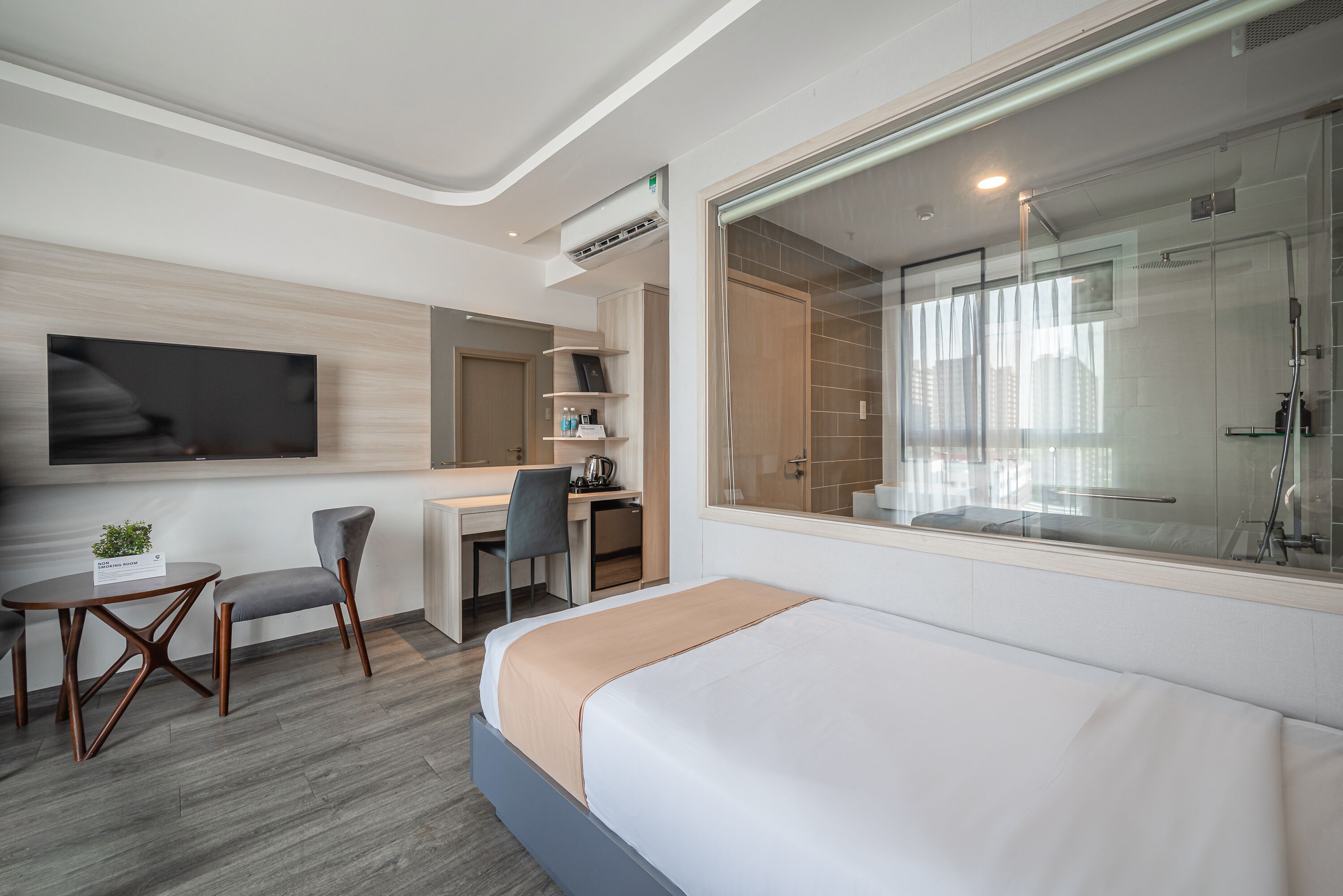 Una habitación de hotel moderna con una cama, un escritorio con una silla, un televisor montado en la pared y un área de ducha con puerta de vidrio.