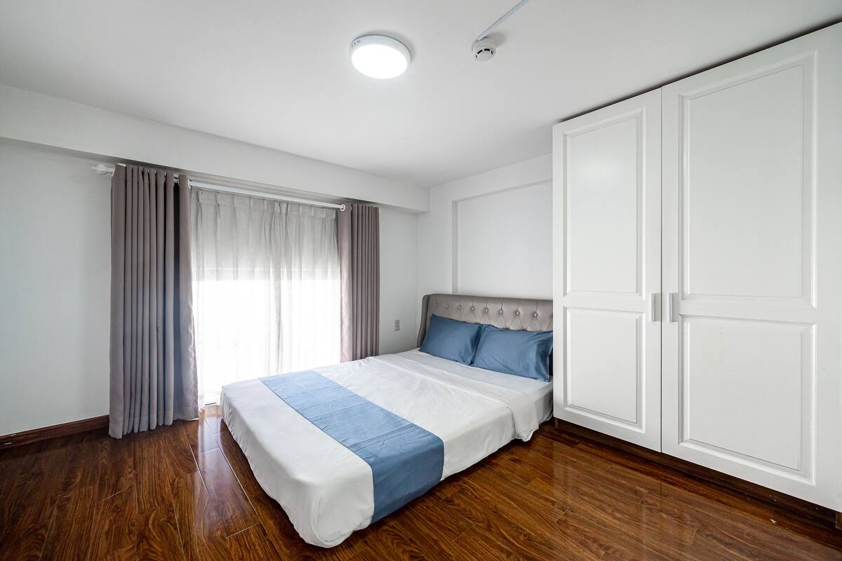 A bedroom with a bed, wooden flooring, a wardrobe, and a window with curtains.