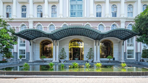 Ein prachtvolles Gebäude mit einer symmetrischen Fassade, einem zentralen Eingang mit dekorativem Bogen und einem umgebenden Wasserspiel mit Pflanzen.