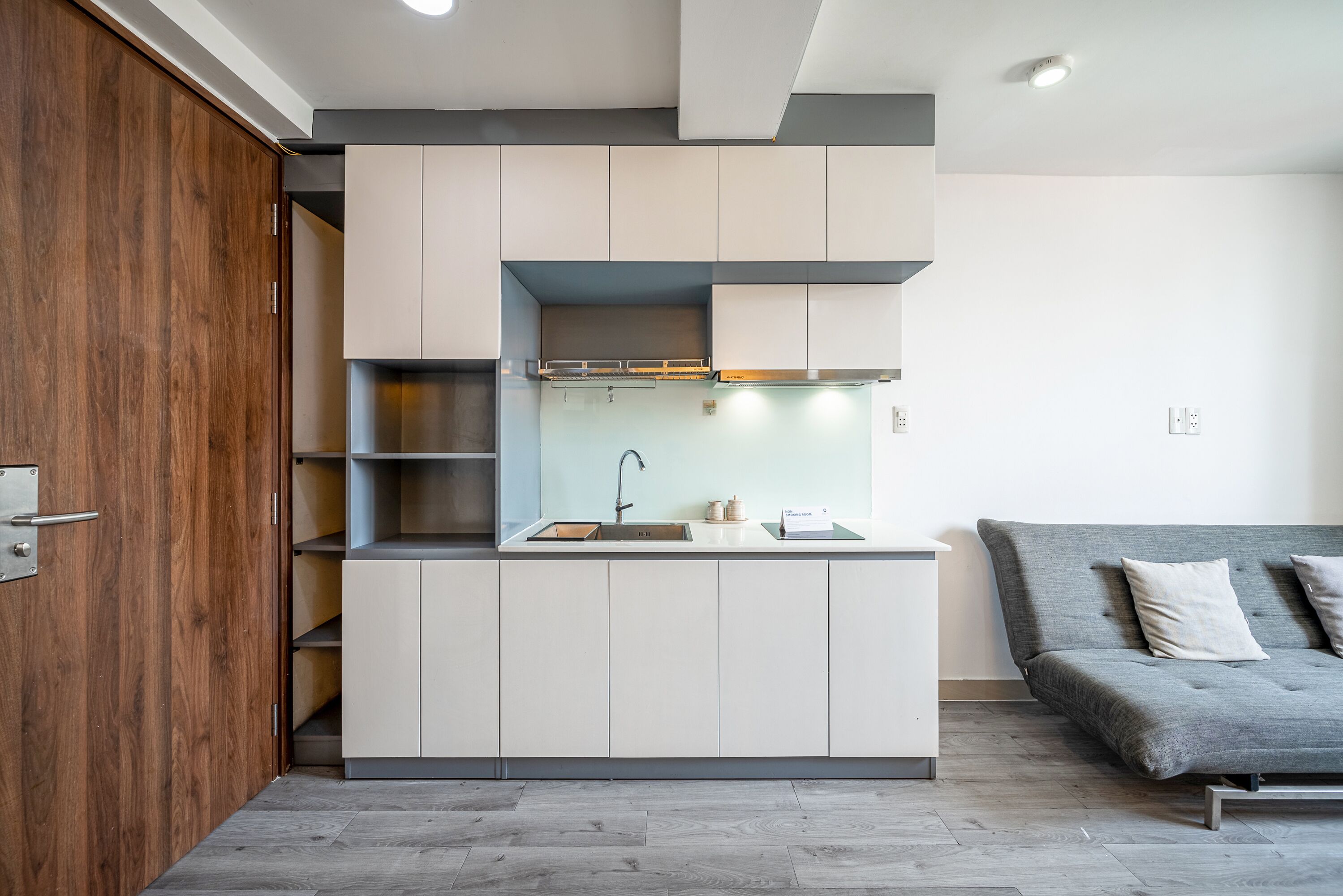 A compact kitchen with white cabinets, a built-in stove, and a grey sofa in the adjacent living area.