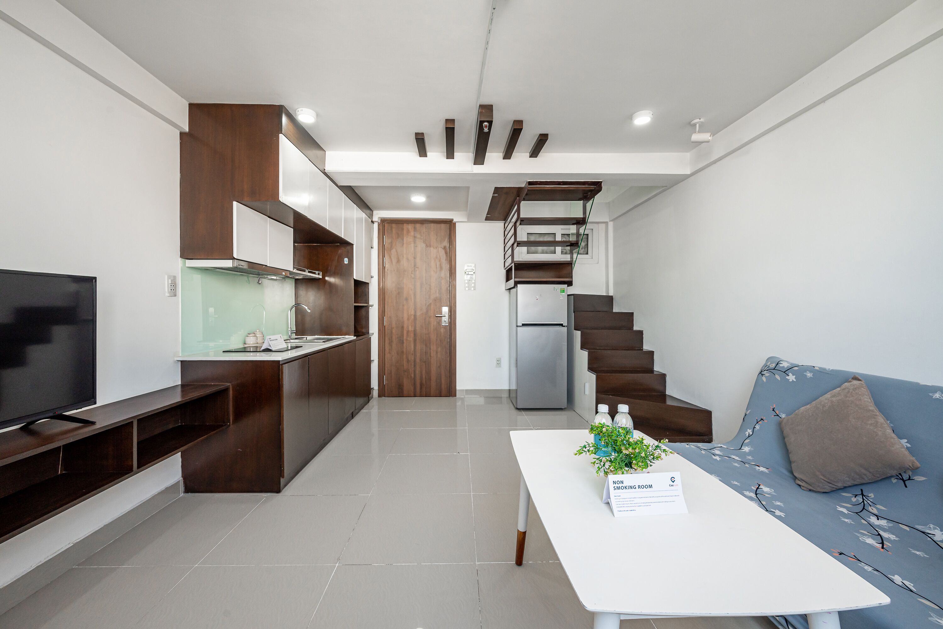 Una cocina moderna con armarios de madera, estufa integrada y refrigerador. Una escalera conduce a un altillo. En la zona de estar, hay una mesa blanca con una planta y un letrero.