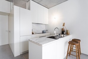 A modern kitchen with white cabinetry, a marble countertop, a stainless steel sink, and wooden stools. - GuestReady - The Modern Silveira (Porto)