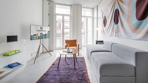 Une chambre d’hôtel moderne avec un canapé gris, un tableau abstrait aux couleurs vives, un tableau blanc portant le logo « GuestReady » et une vue sur la ville par la fenêtre.
