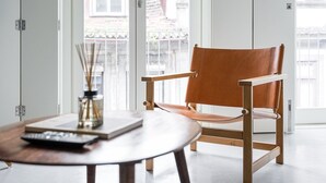 Une chambre moderne avec une chaise en bois, une table sur laquelle se trouvent un livre et une télécommande, et de grandes fenêtres donnant sur les bâtiments environnants.