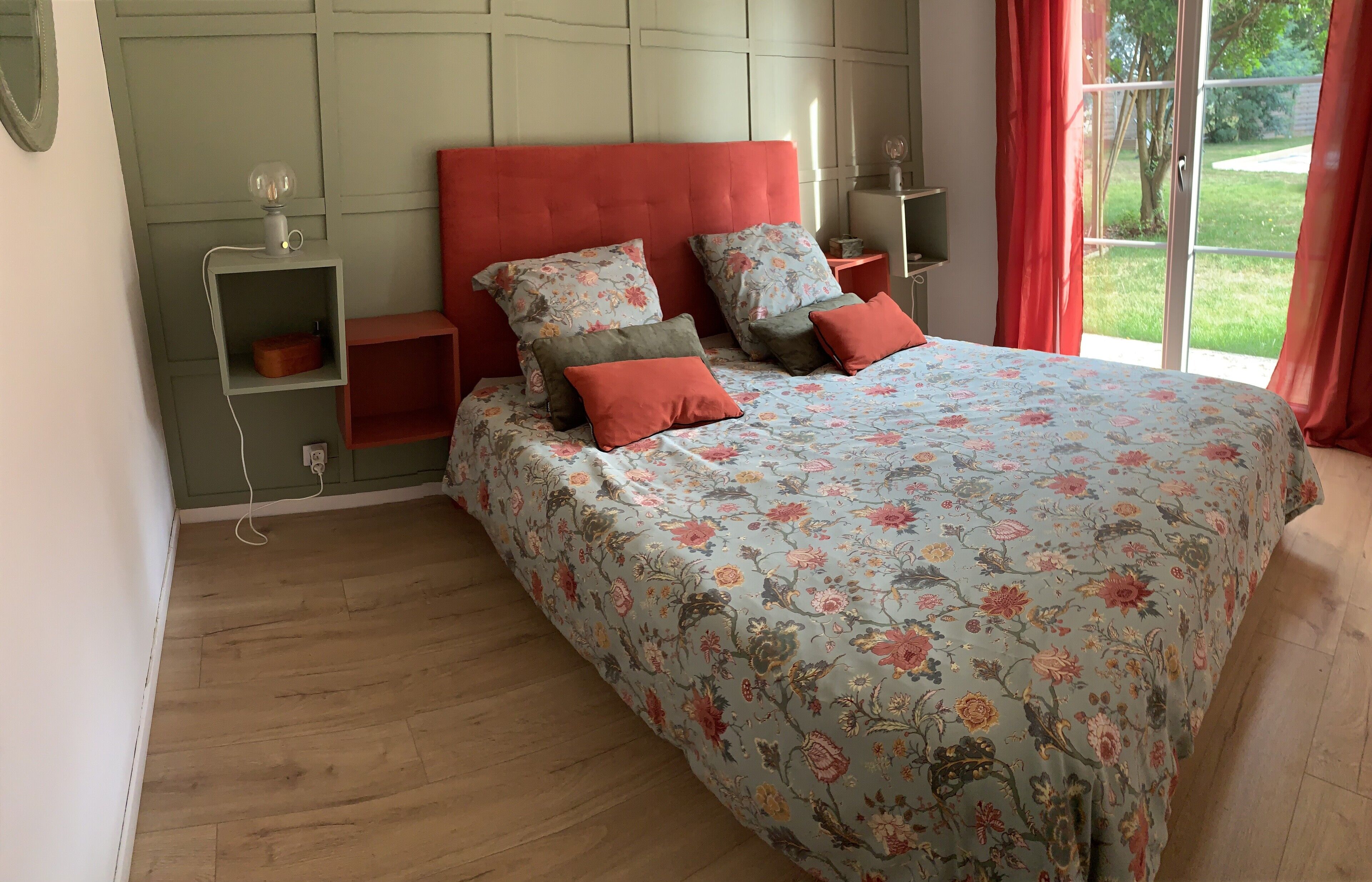 A bedroom with a floral bedspread, red pillows, a red headboard, and a view of a garden through a sliding door.