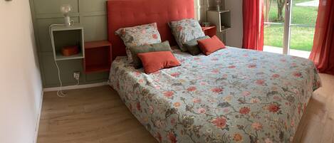 A bedroom with a floral bedspread, red pillows, a red headboard, and a view of a garden through a sliding door.