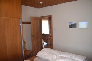A bedroom with a wooden wardrobe, a bed with white bedding, a wooden chair, and two framed pictures on the wall.