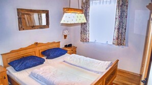 Un dormitorio con cama de madera, dos almohadas, una mesita de noche, una lámpara de pared, un espejo y una ventana con cortinas florales.