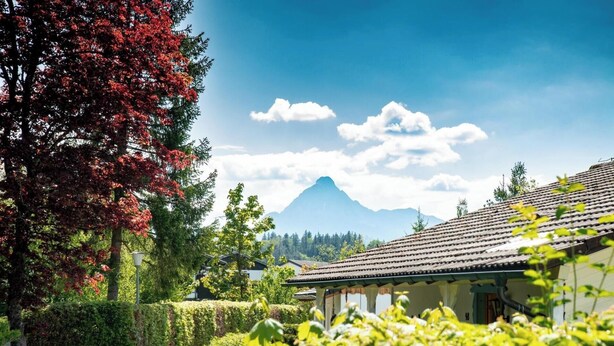 A pathway leading to a traditional building with a tiled roof, surrounded by lush greenery and a mountain in the background.