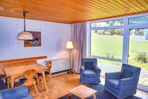 Une pièce avec un plafond en bois, une table à manger avec des chaises, un canapé, une lampe et une vue sur un espace verdoyant grâce à de grandes fenêtres.
