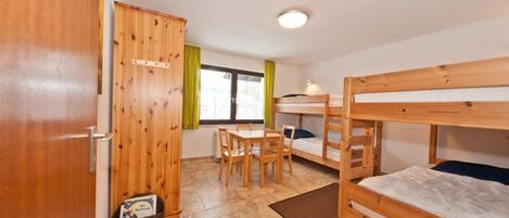 A room with wooden bunk beds, a wooden table with chairs, a window with curtains, and a door.