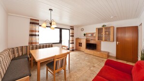 Uma sala de estar com sofá, mesa de jantar, um sofá vermelho e um espaço de cozinha com armários e televisão.