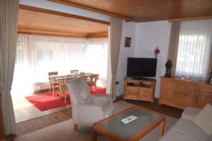 Ein Wohnzimmer mit Fernseher, Holzmöbeln, einer Essecke mit rotem Teppich und großen Fenstern mit Vorhängen.