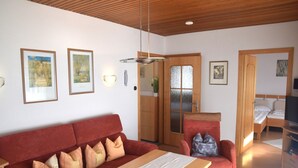 Une chambre moderne avec un canapé rouge, une table en bois, un fauteuil rouge et un lit. Des tableaux encadrés ornent le mur, et un téléviseur est posé sur un meuble.