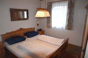 Ein Schlafzimmer mit einem hölzernen Bett, einem Nachttisch, einer Wandlampe, einem Fenster mit floralem Vorhang und einem Spiegel.