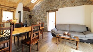 A cozy living room with a stone wall, wooden ceiling beams, a grey sofa, and a wooden dining table with chairs.