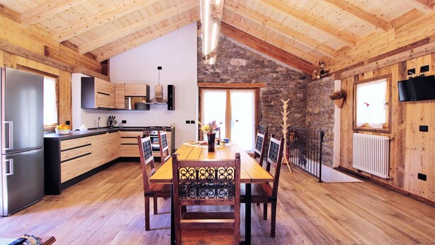 A wooden interior with a dining table, chairs, a kitchen area, and a stone wall.