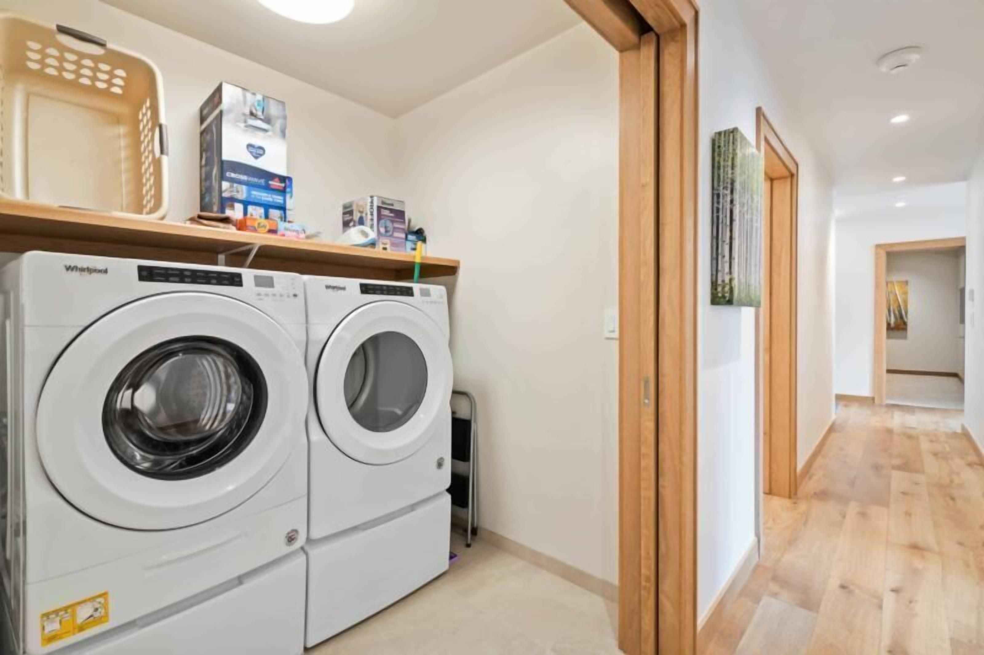 A laundry room with two Whirlpool washers, a wooden shelf with various items, and a hallway leading to another room.