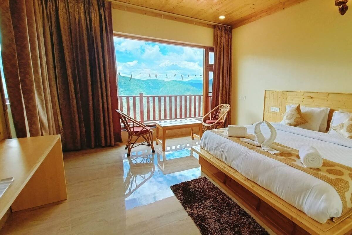 Quarto com cama e escrivaninha de madeira, uma cadeira e varanda com vista para as montanhas.