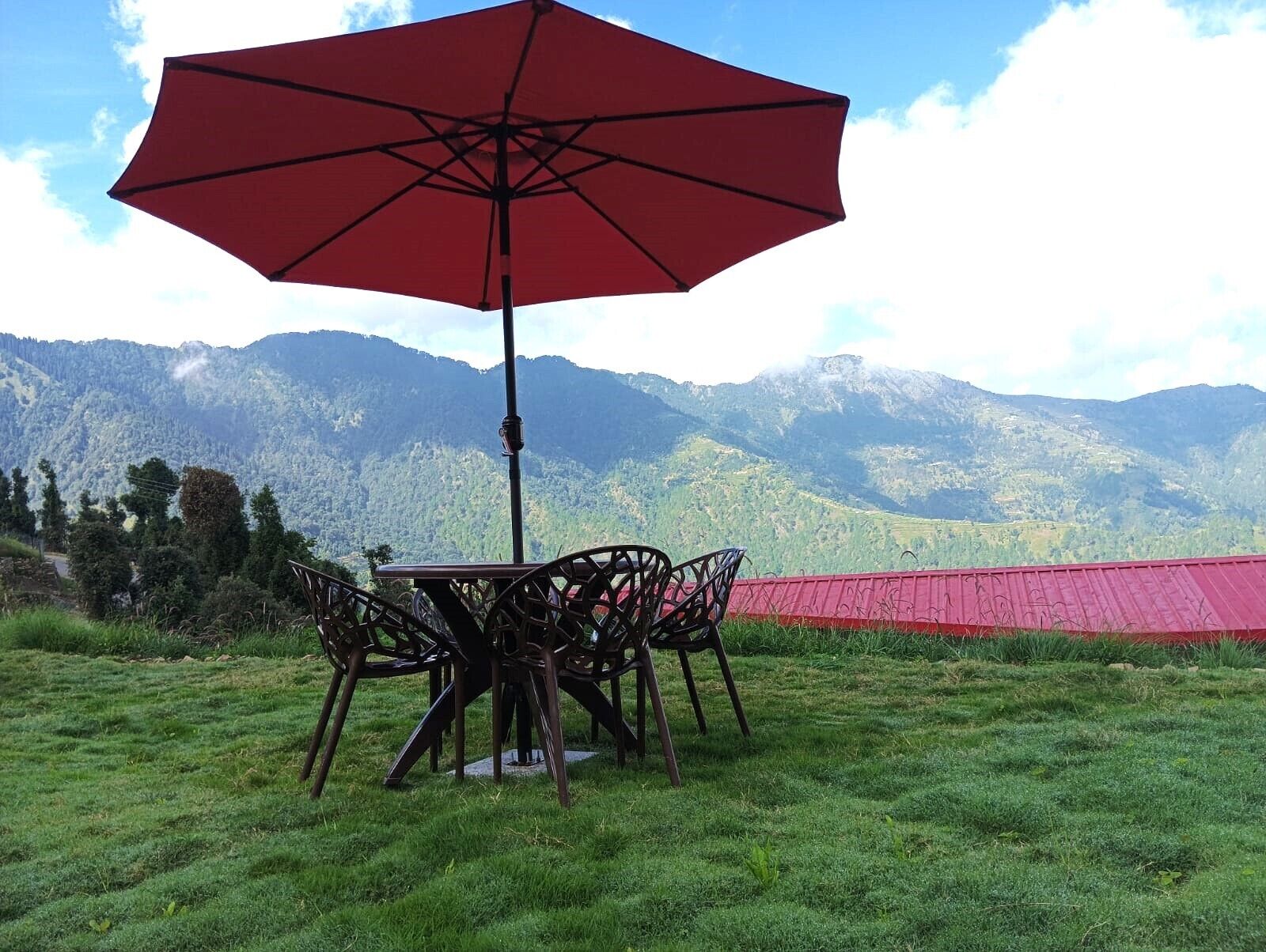 A table with chairs and a red umbrella in a grassy area with a mountain backdrop.