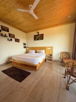 A wooden-framed bedroom with a bed, a ceiling fan, a painting, and a dining area with wicker furniture.