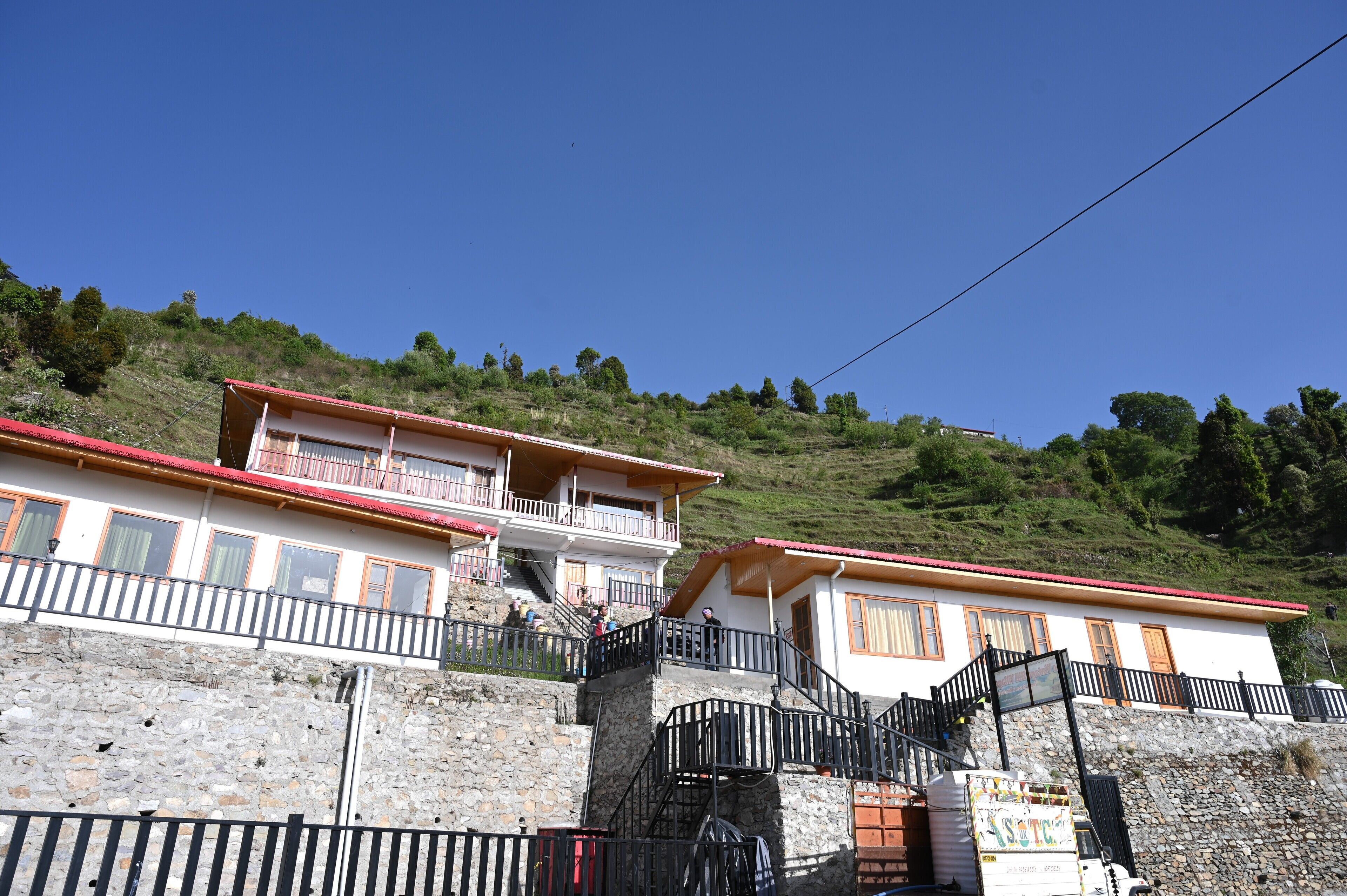 A two-story building with a red roof, white walls, and balconies. There are stairs leading up to the building, and a stone wall in the foreground.