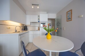 A modern kitchen with white cabinets, a white dining table with a yellow vase, and blue chairs. - Well-maintained apartment with sea view (Middelkerke)