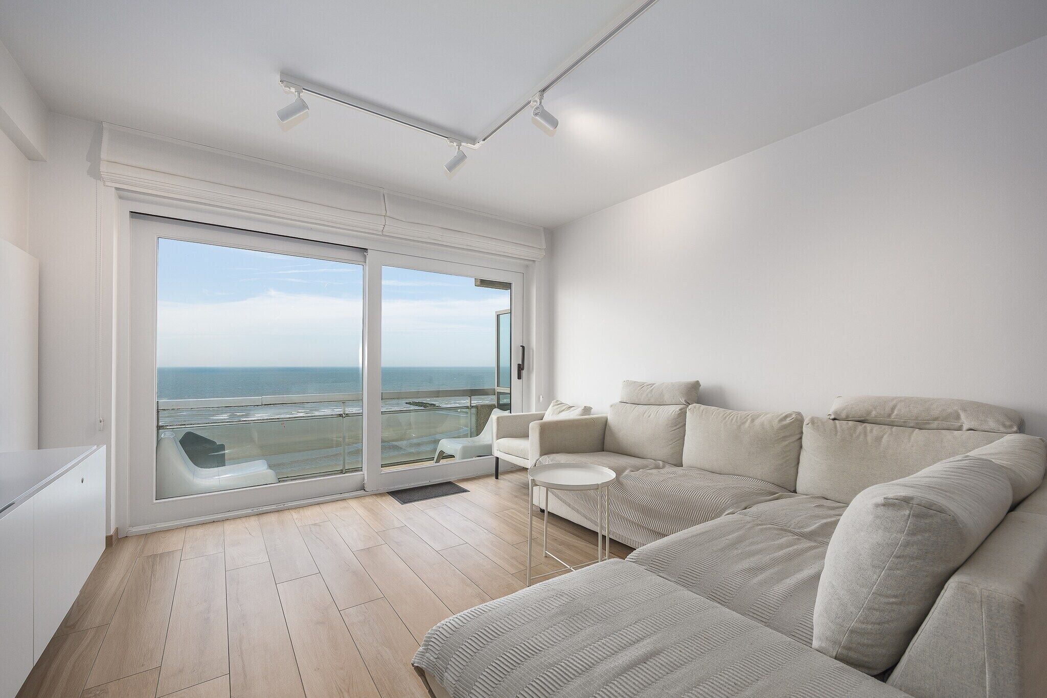 A modern living room with a large sliding glass door to a balcony, a beige sofa, a small white side table, and a white cabinet.