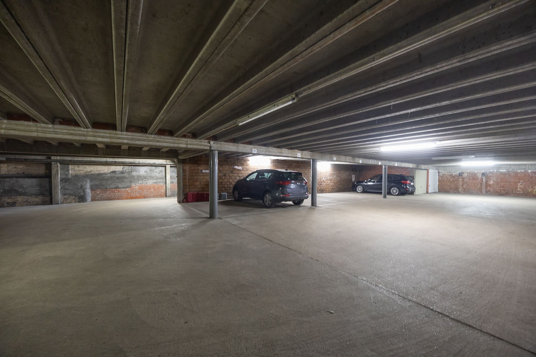 En parkeringskælder med betongulv og murstensvægge, der viser flere parkerede biler og lysarmaturer i loftet.