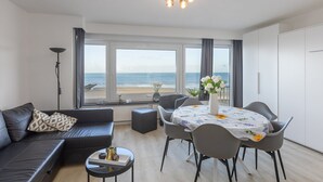 A modern living room with a sofa, dining table, and chairs, large windows with a view of the beach, and a ceiling light fixture. - Cosy studio with sleeping area and sea view (Middelkerke)
