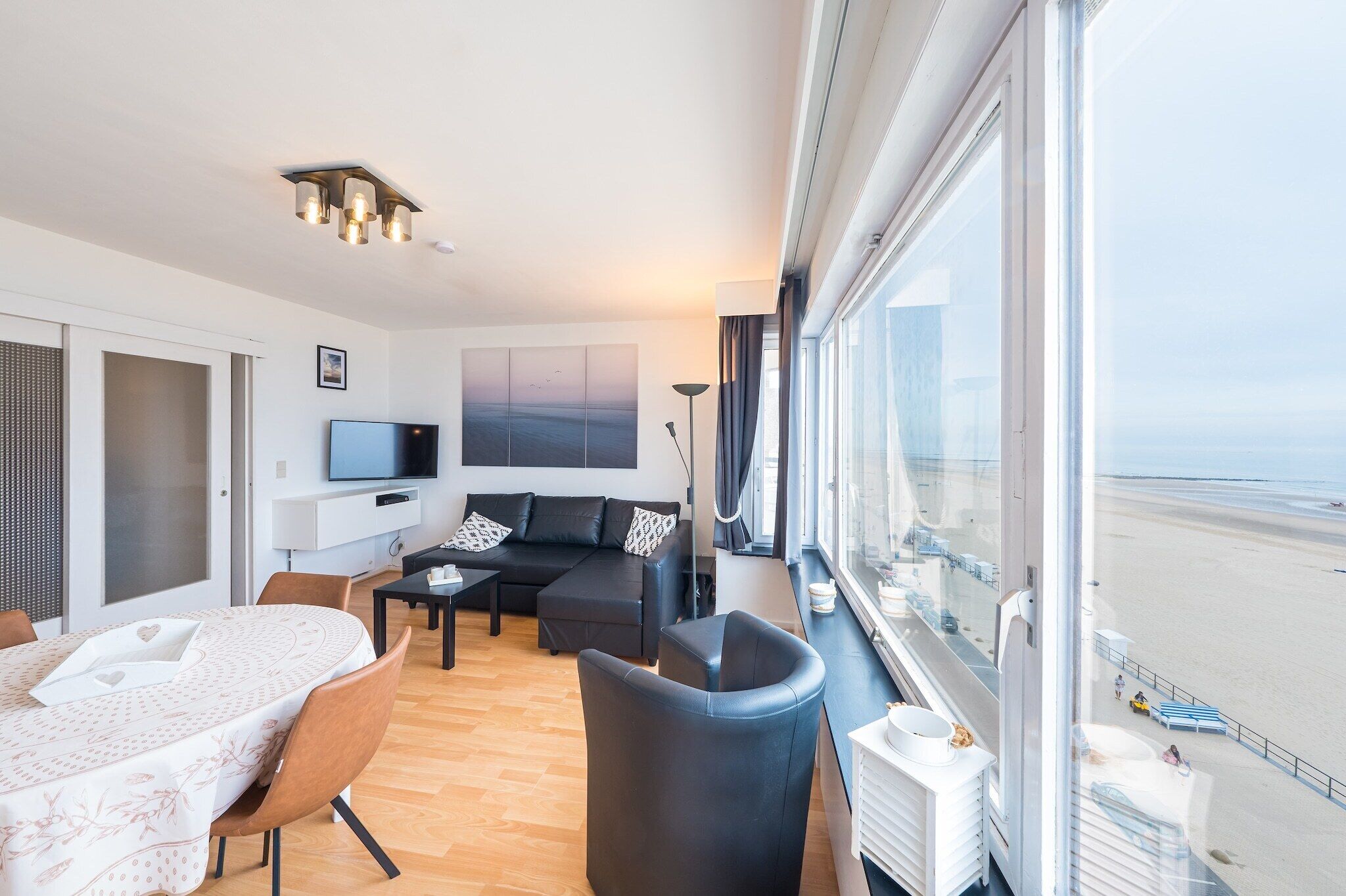 A modern hotel room with a large window, a sofa, a dining table, and a TV.