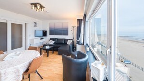 A modern hotel room with a large window, a sofa, a dining table, and a TV. - Cosy studio with sleeping area and sea view (Middelkerke)