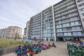 A modern multi-story building with a flat facade, large windows, and a paved area in front with children's ride-on toys. - Beautiful apartment with phenomenal sea view (Westende)